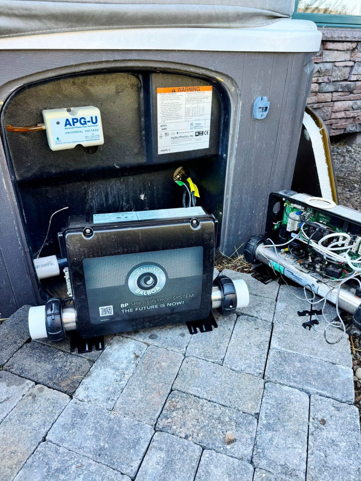 Open hot tub equipment bay showing internal wiring and control system components on stone pavers