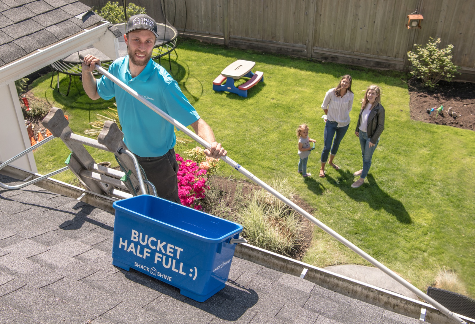 A uniformed Shack Shine technician working safely at roof level with a smiling family in the backyard below