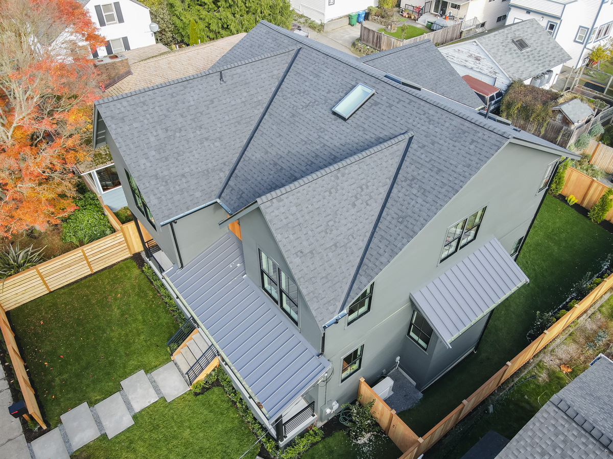 High-angle comprehensive roof view showing charcoal shingles, ridge cap shingles, skylight, and standing seam metal awnings
