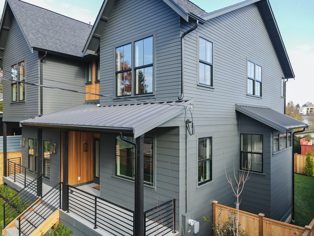 Side and front angled view of standing seam metal roofing, downspouts, dark trim windows, and cedar wood fence