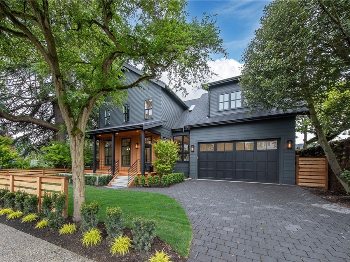 Full front elevation showing dark garage, stone paver driveway, mature shade tree, and cabin-style metal roof porch