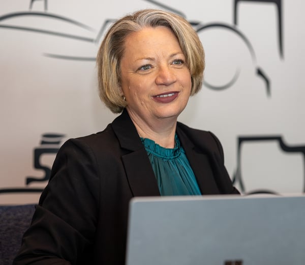 A smiling woman with short blonde hair in a dark blazer sits at a desk with a laptop.