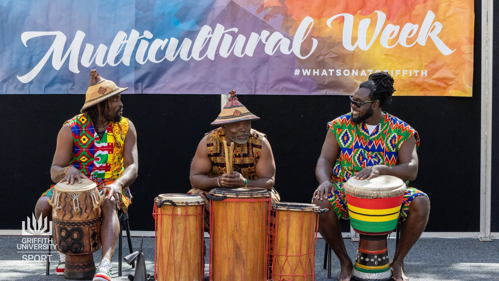 African Beat master drummers in vibrant kente prints and traditional woven hats smiling behind acoustic djembe and dunun drums during Multicultural Week