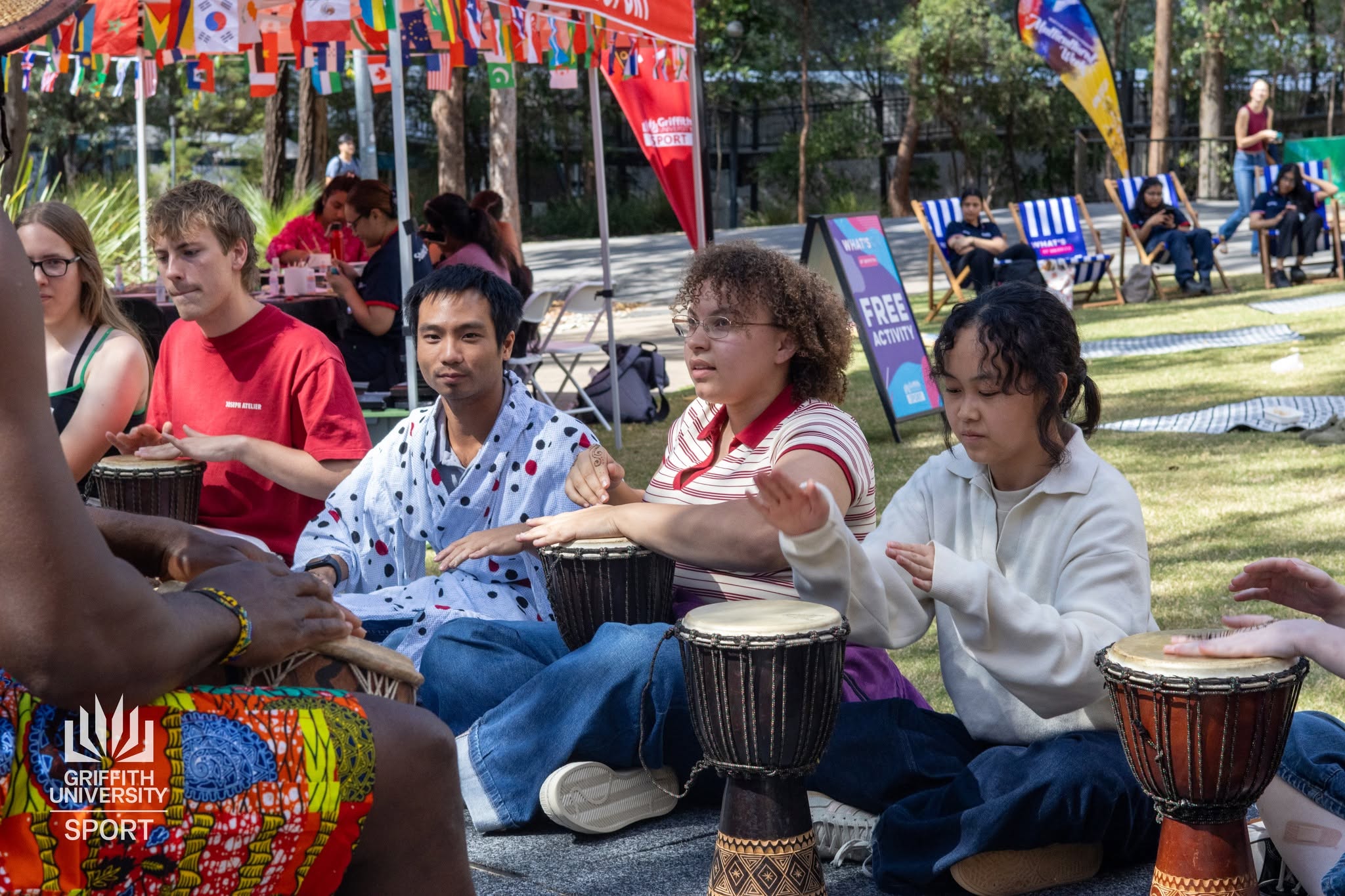 University students seated on a campus lawn participating in a hands-on djembe drum circle