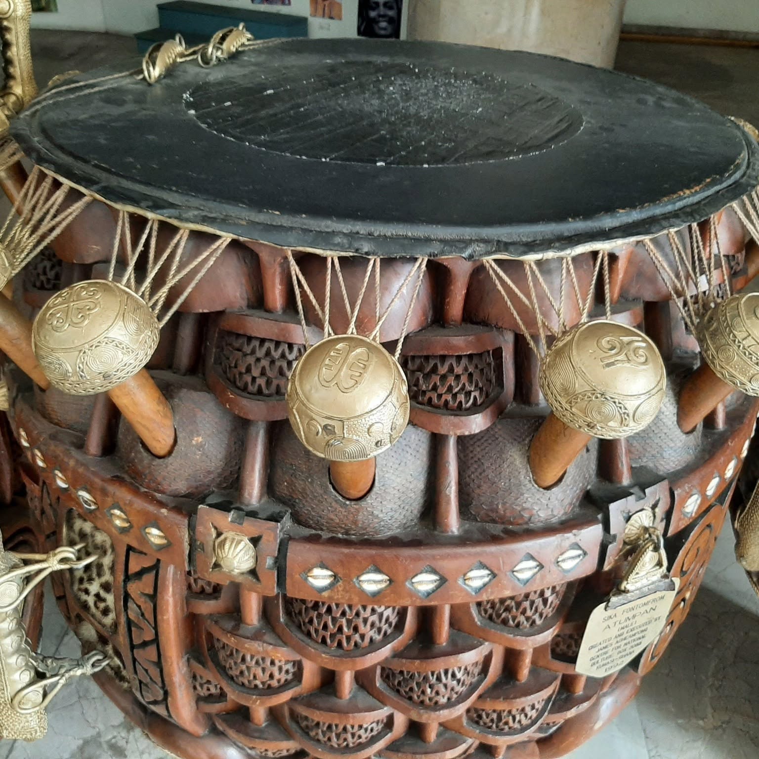 Close-up of a royal ceremonial djembe drum head with ornate gold-engraved tuning pegs, cowrie shell inlays, and woven leather wrap