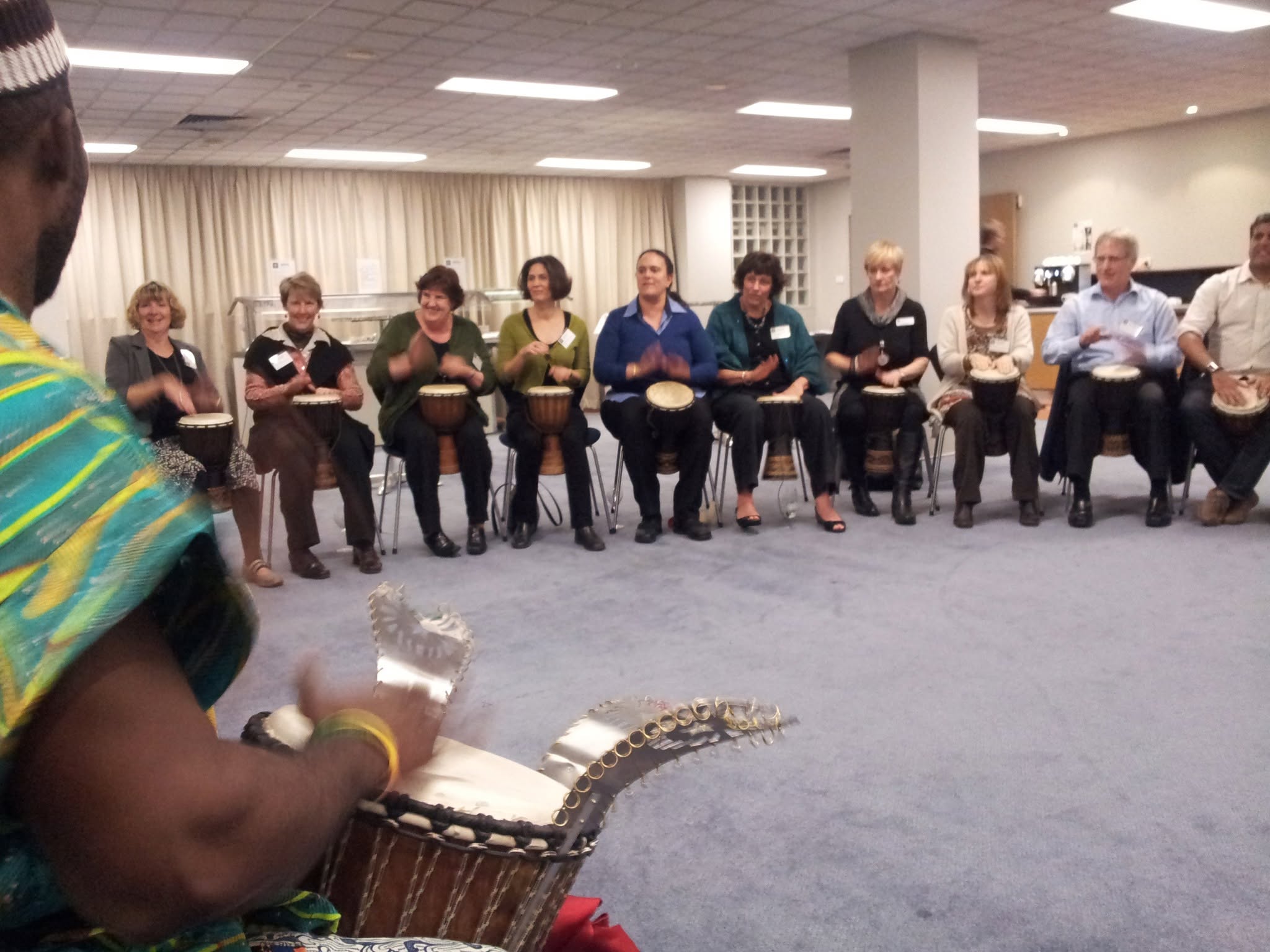 Adult professionals seated in a wide boardroom circle with djembes being led by an African Beat master drummer in traditional attire