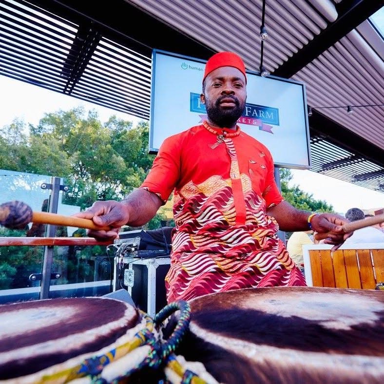 Performer in a red traditional patterned shirt drumming with mallets on an outdoor campus stage setup