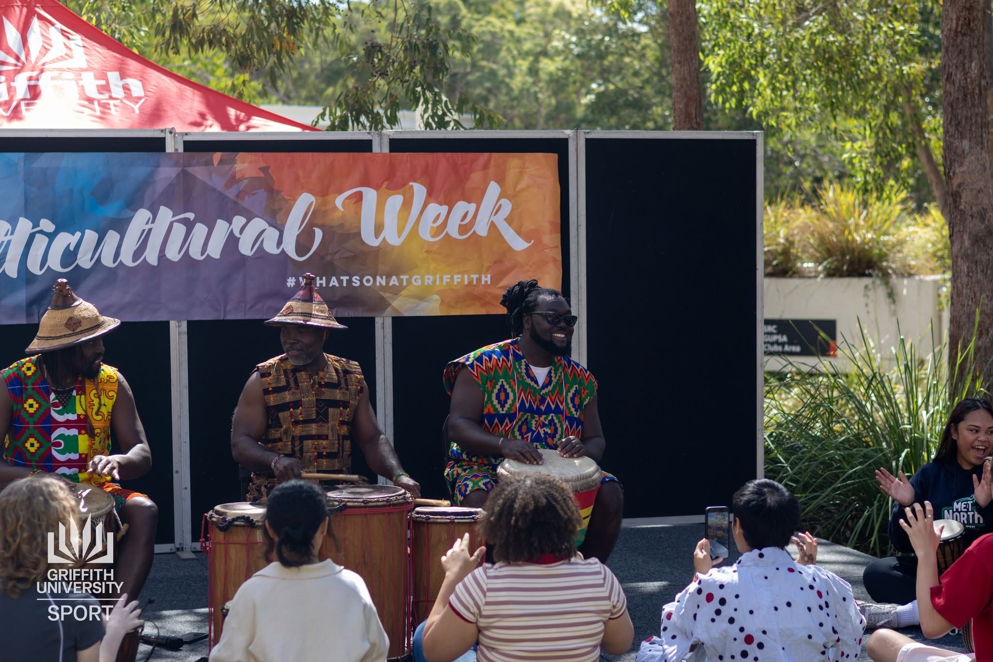 African Beat African drummers in traditional woven attire performing on stage beneath a Multicultural Week banner with an engaged student audience
