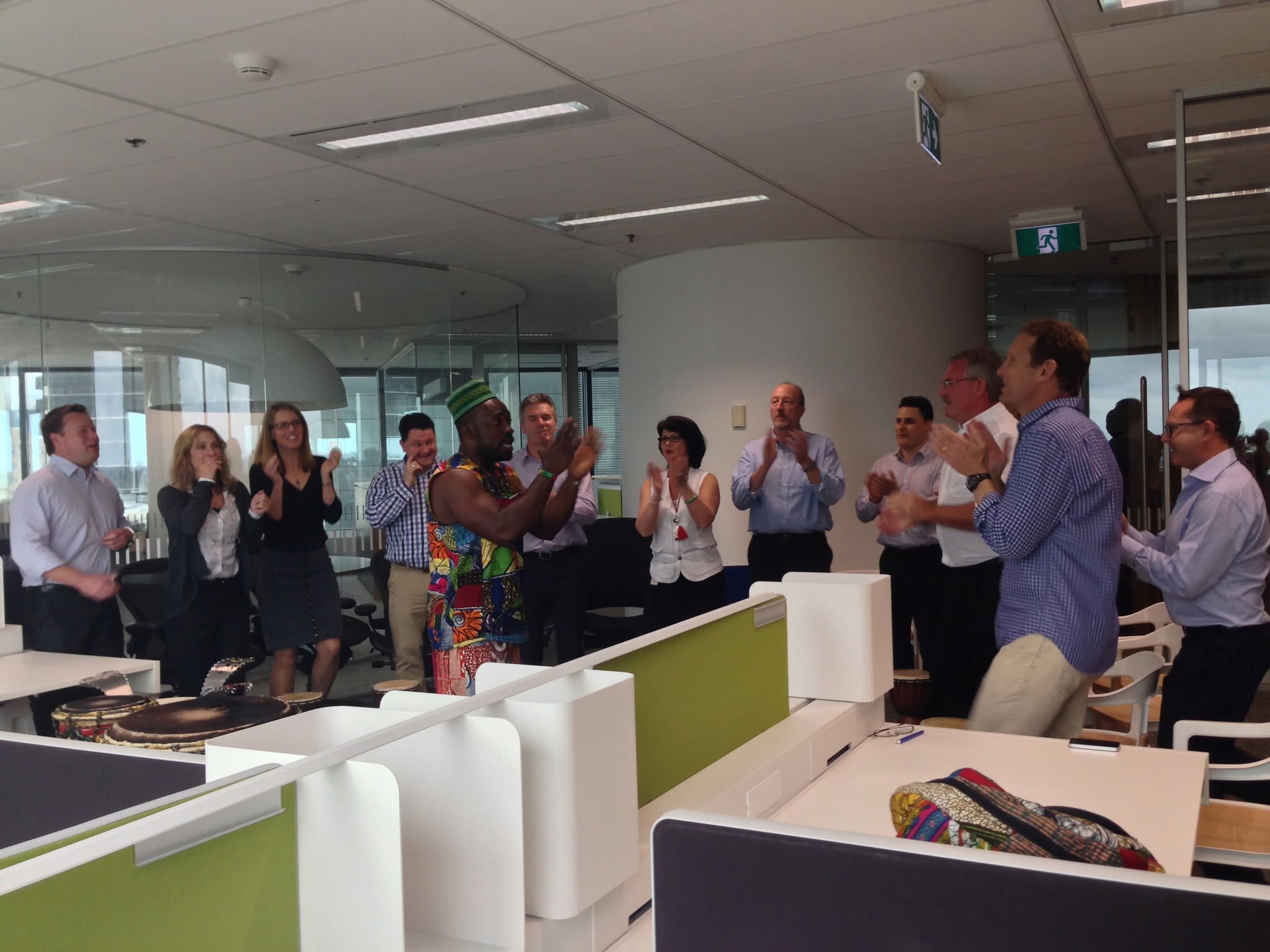 Office team standing and clapping enthusiastically among open-plan desks alongside an African Beat lead drummer in vibrant print attire