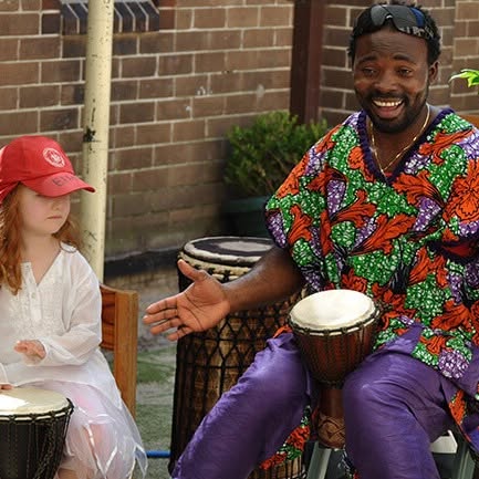 African Beat lead performer in vibrant kente-style dashiki smiling warmly with a djembe drum while teaching a young participant