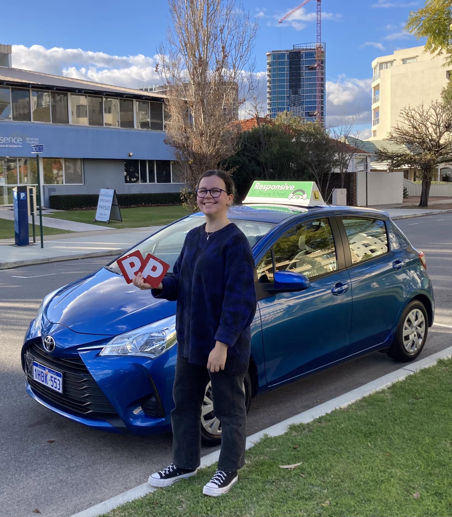 Smiling student with glasses holding two red P plates beside a blue Toyota Yaris