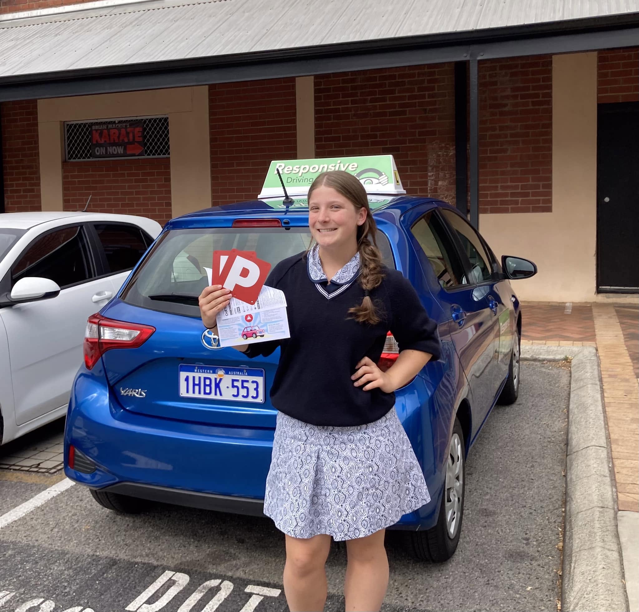 Smiling student in school uniform holding red P plates and a leaflet in front of a blue Toyota Yaris