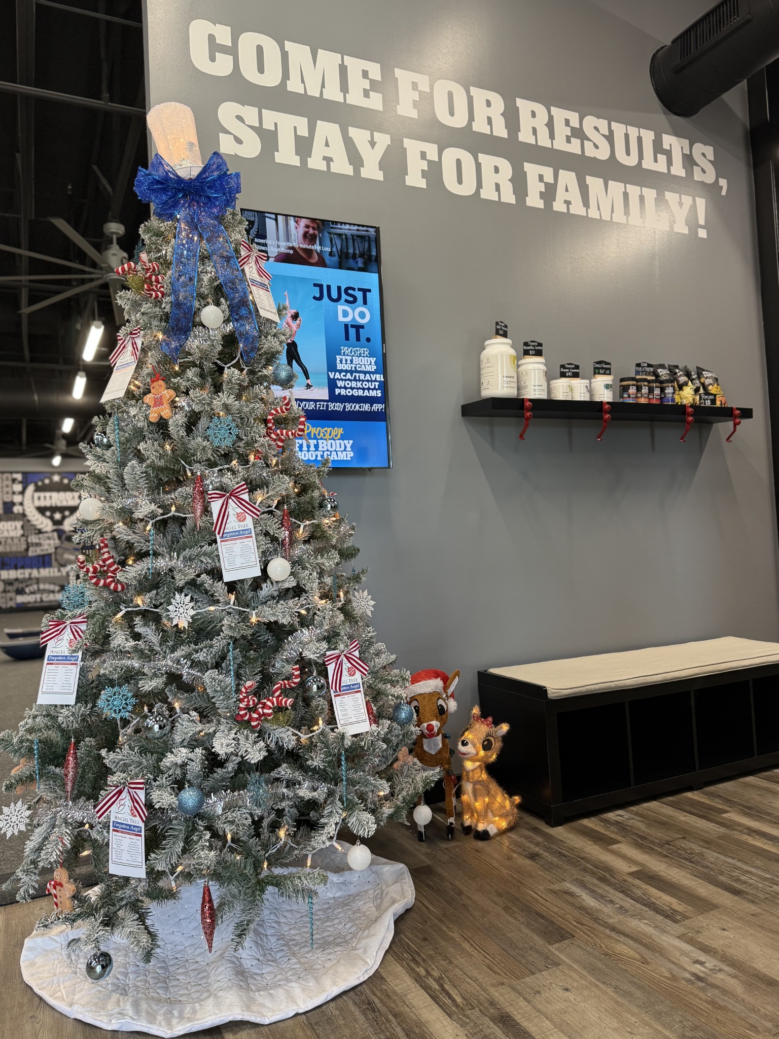 Authentic Prosper Fit Body Boot Camp Angel Tree decorated with blue bow, hanging Angel Tree tags, and Come For Results Stay For Family wall lettering