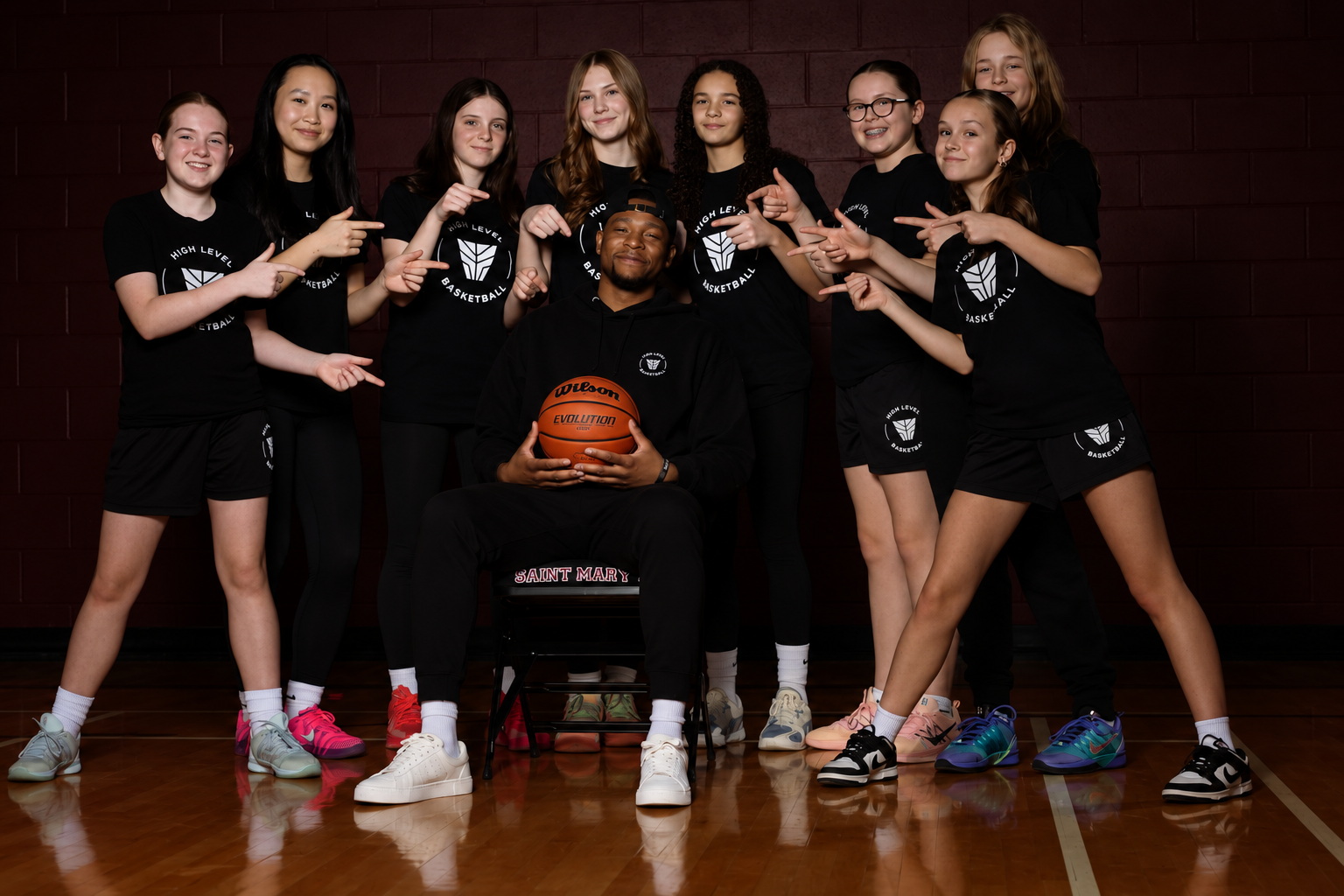 A coach seated with basketball surrounded by smiling youth basketball athletes in High Level shirts pointing toward him