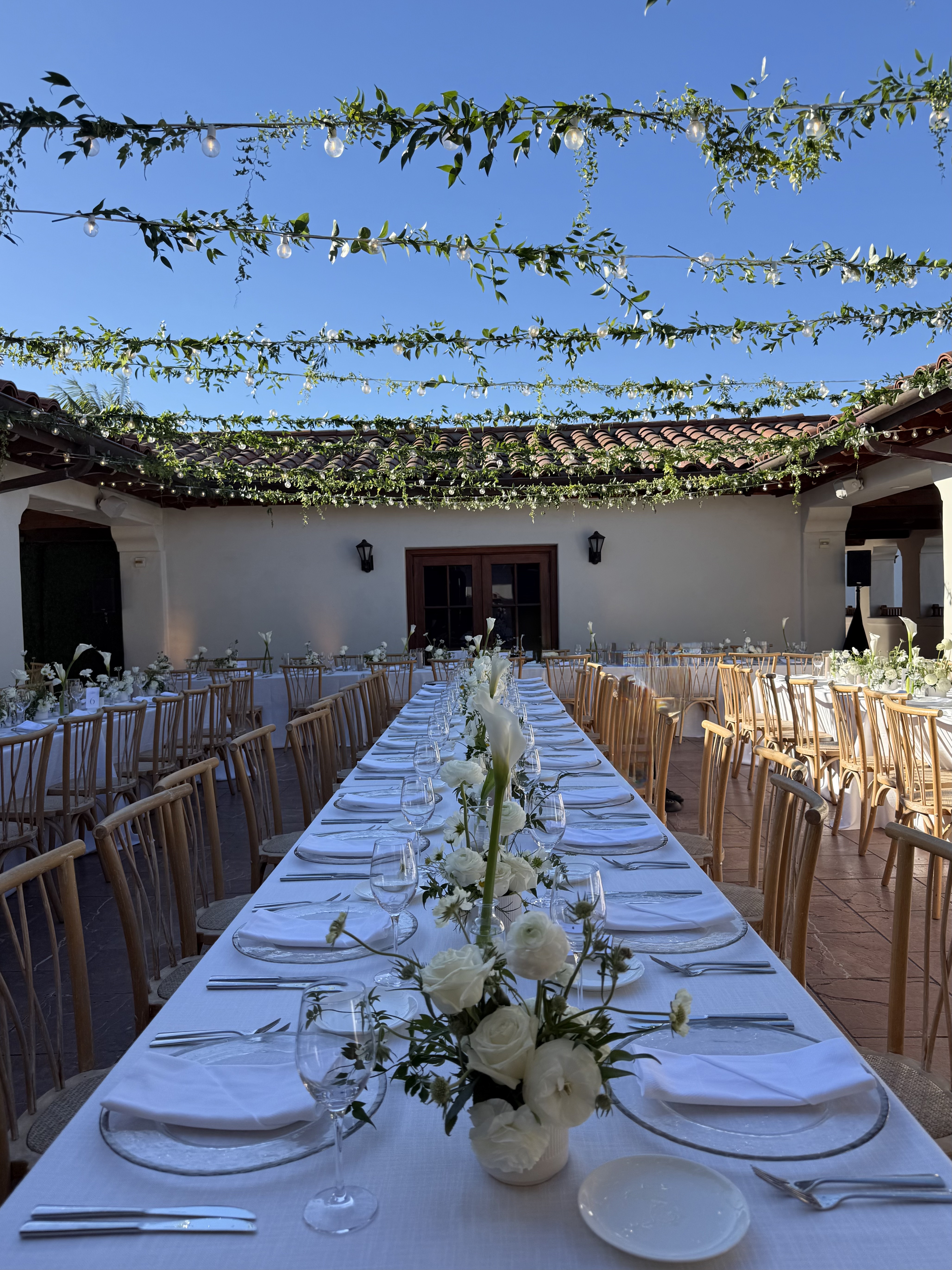 Long courtyard dinner tablescape with white florals and overhead string lights by Soulform Florals, Los Angeles