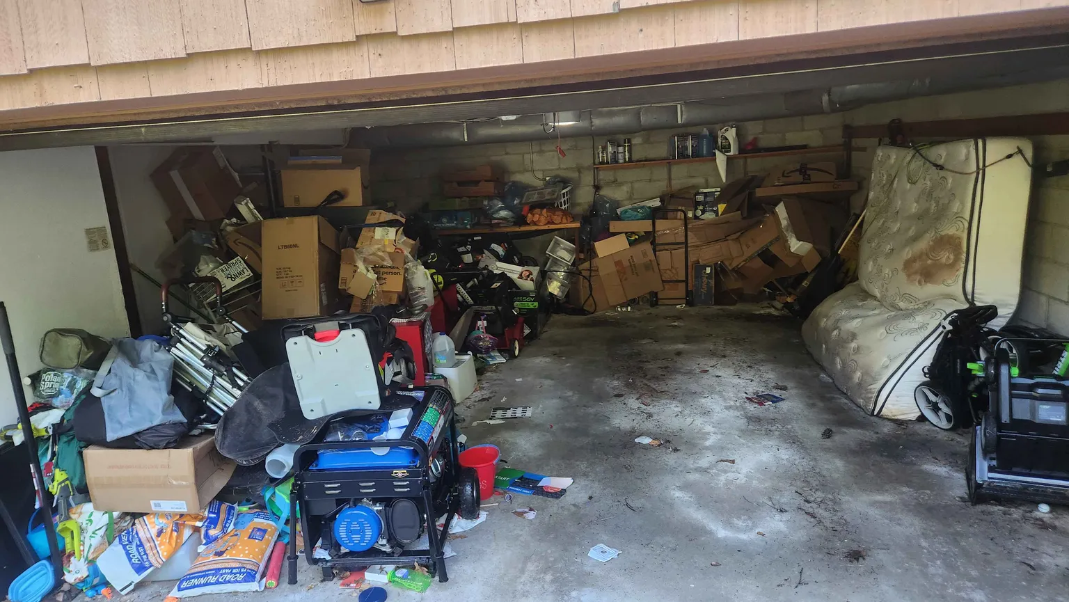 Cluttered garage with stained mattress, power equipment, and piles of cardboard boxes before cleanout