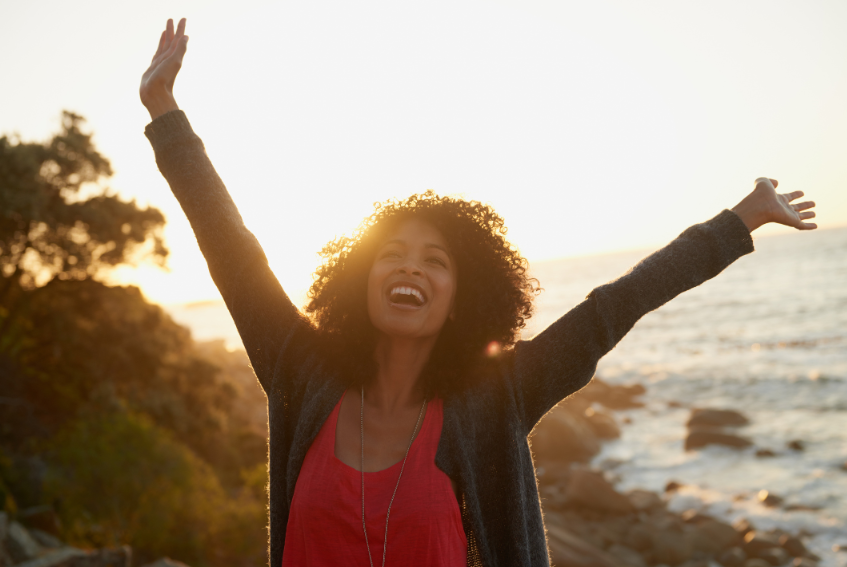 Happy radiant woman celebrating with arms raised in warm coastal golden sunlight
