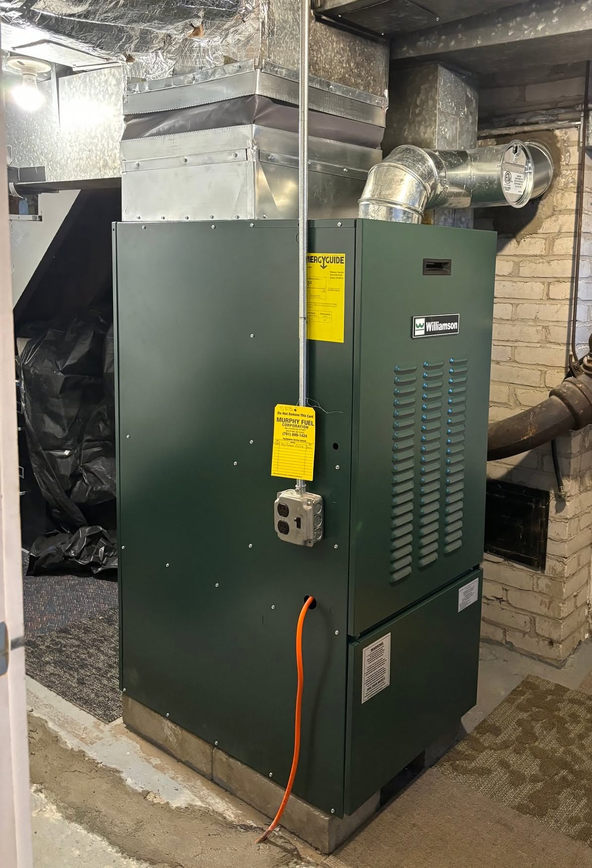 Forest green Williamson residential warm air oil furnace installed on elevated concrete pad with custom galvanized supply air transition plenum, vibration canvas collar, electrical service disconnect, yellow EnergyGuide label, and official heating service inspection tag