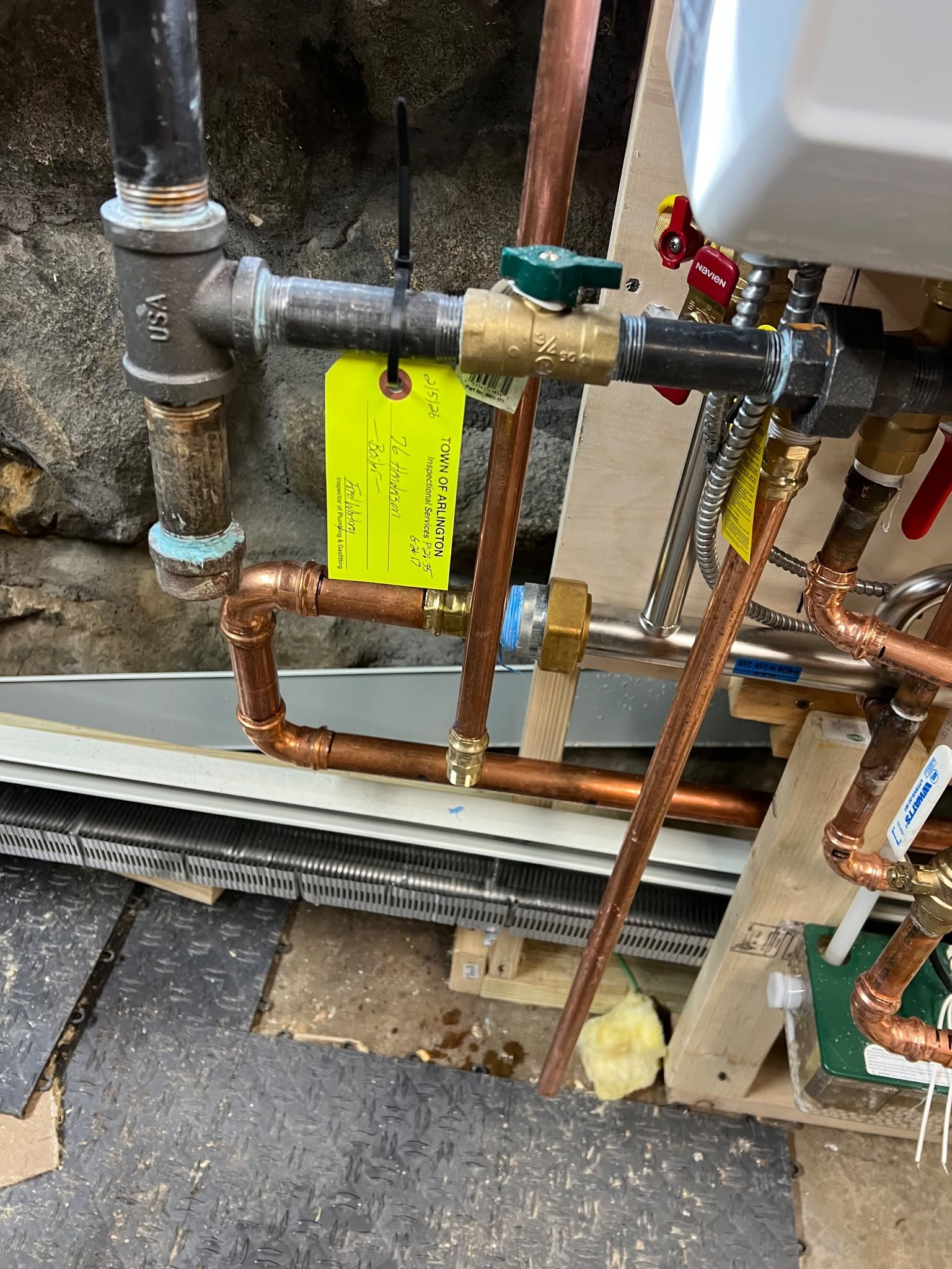 Close-up of municipal plumbing and heating inspection approval tag signed by licensed inspector on black-iron gas pipe manifold and copper boiler piping