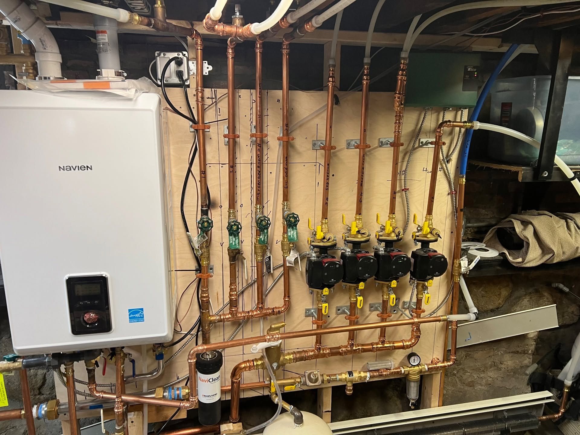 Complete 5-zone hydronic manifold on finished plywood board with Grundfos circulator pumps, Navien combi boiler, NaviClean magnetic filter, and copper distribution piping