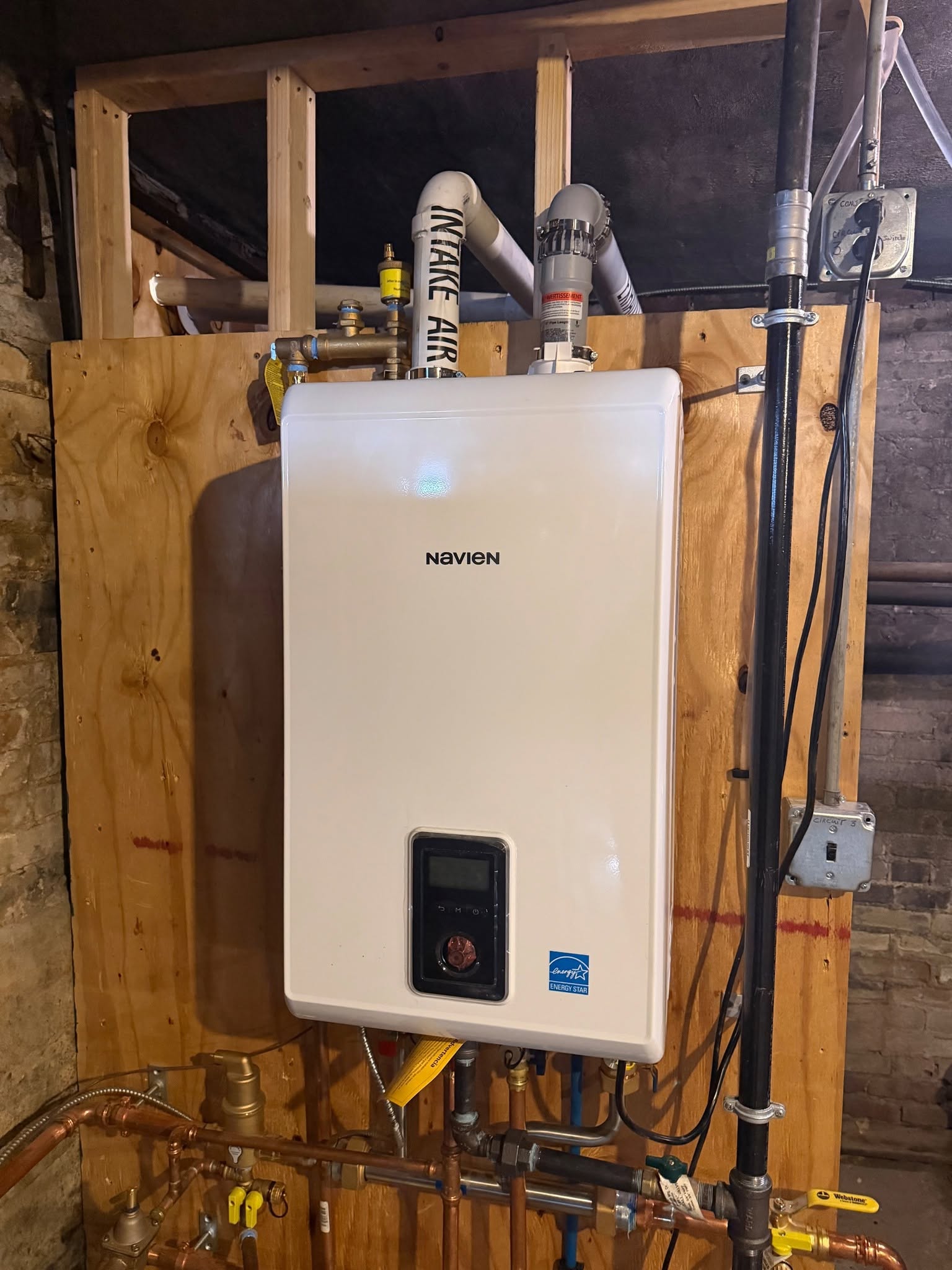 Wall-mounted white Navien condensing heating unit on plywood backing with dedicated PVC intake air pipe, exhaust flue, gas line sediment trap, and electrical emergency shutoff switch