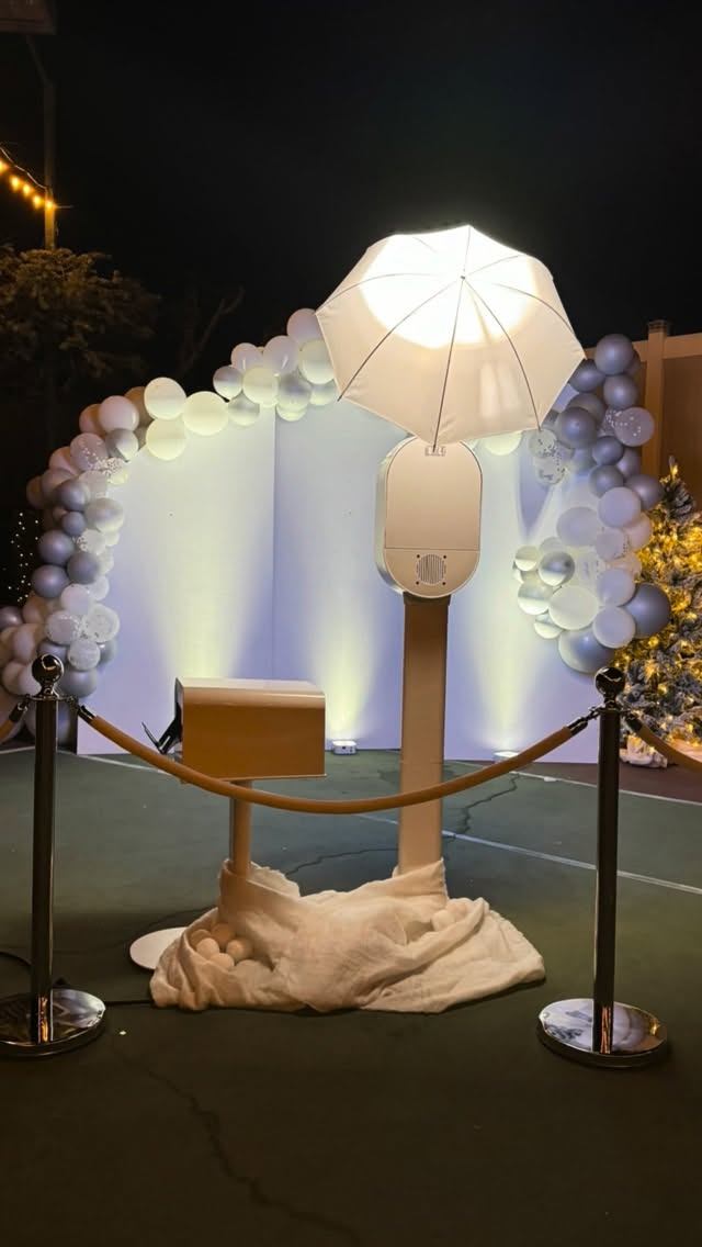Outdoor evening celebration setup with white photo booth, umbrella strobe, and silver-white balloon arch