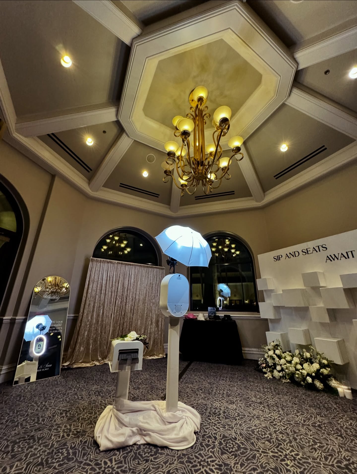 Luxury ballroom rotunda setup with white photo booth, umbrella light, champagne drapes, and chandelier