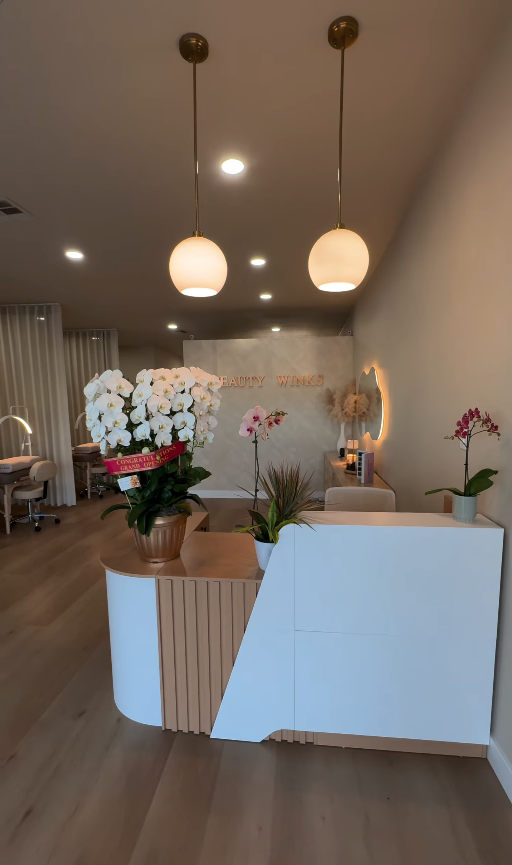 Inside the new Beauty Winks studio reception on N Tustin Ave, featuring the white and fluted oak front desk, gold lettering, fresh white orchids, and frosted globe pendant lights