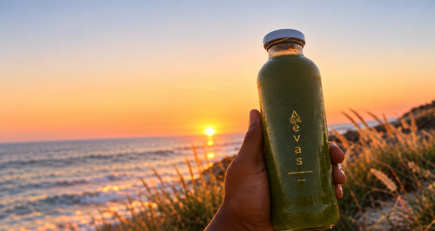Hand holding an authentic 16 oz green AEVAS cold-pressed juice bottle against a Pacific coastal sunset