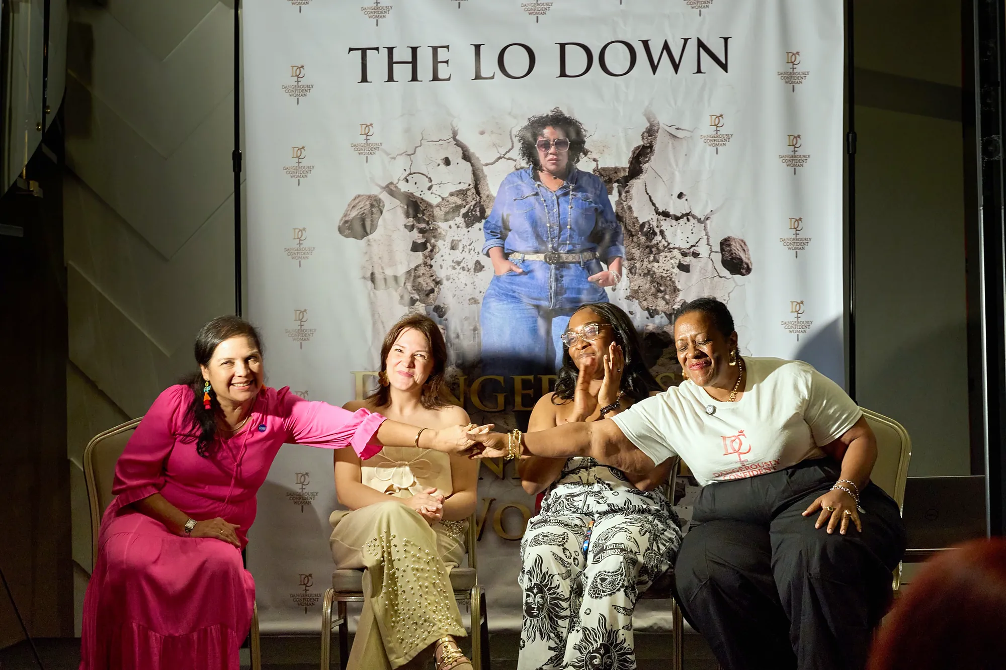Four women smiling and joining hands on stage at a Dangerously Confident Woman event hosted by The Lo Down with Isha Lo