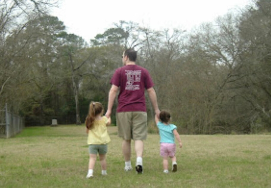 Rachel's husband walking in the grass holding their two young daughters' hands
