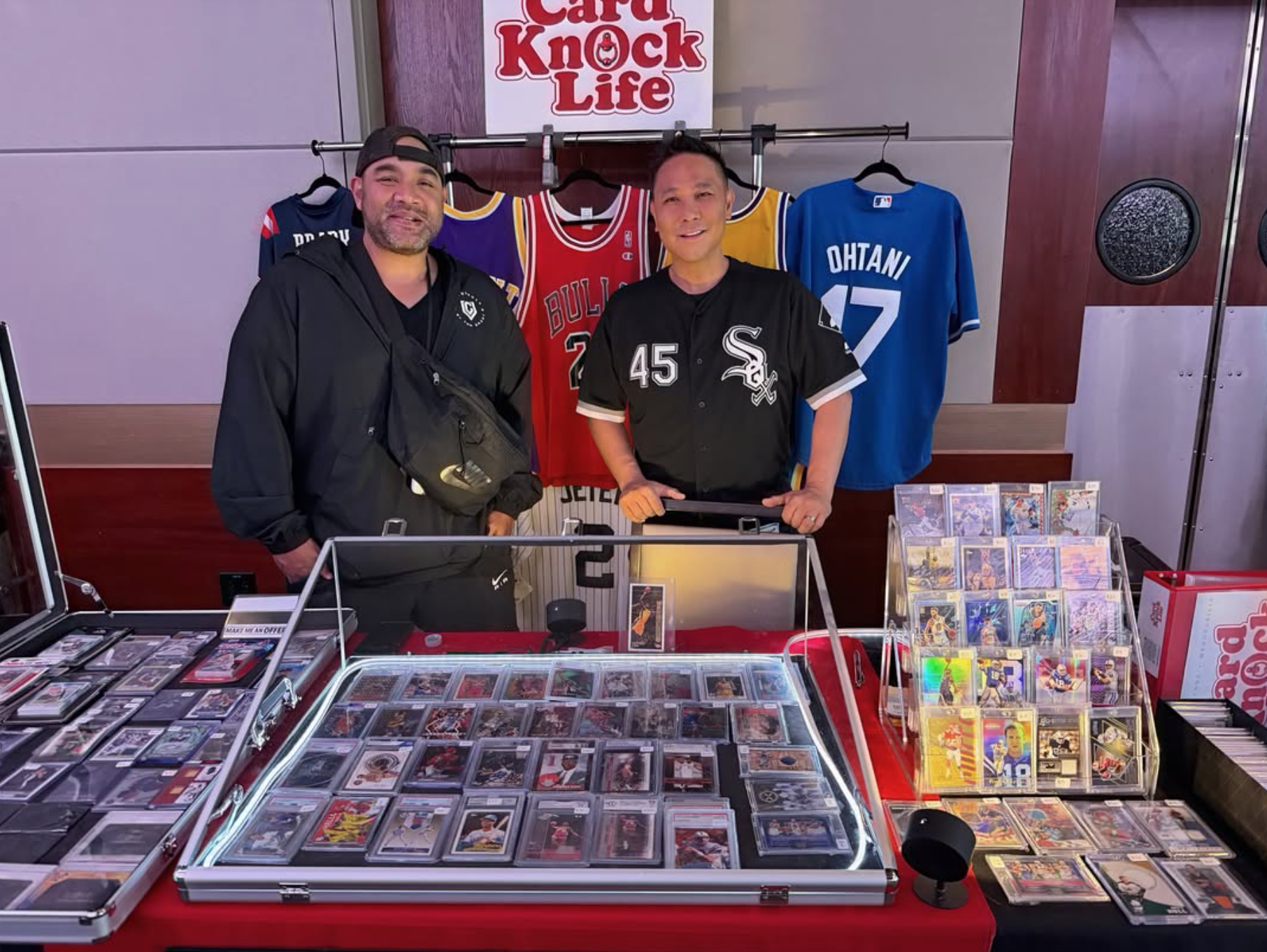 Card Knock Life TX crew behind their loaded sports cards display table with graded slabs and jerseys at a Texas card show