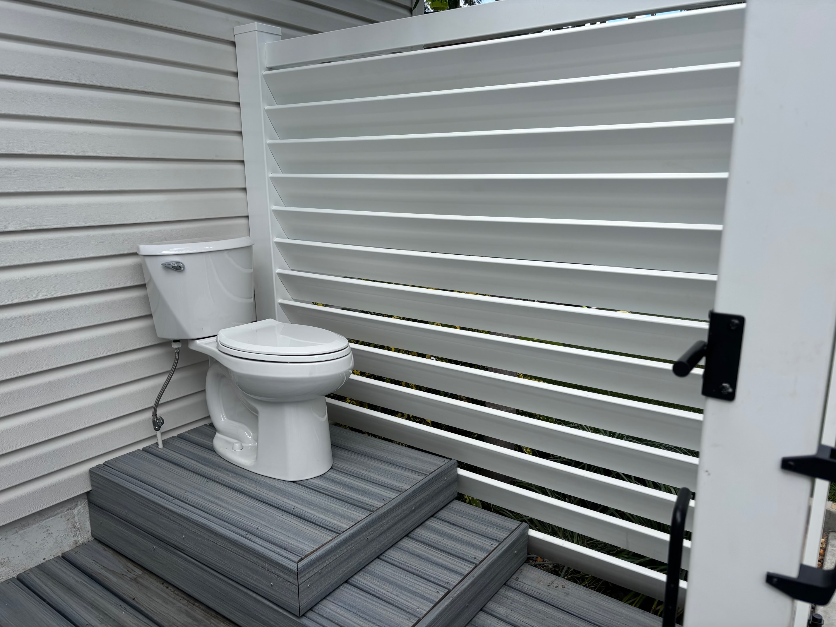 Outdoor toilet mounted on two-tiered grey composite decking next to a white louvered privacy partition