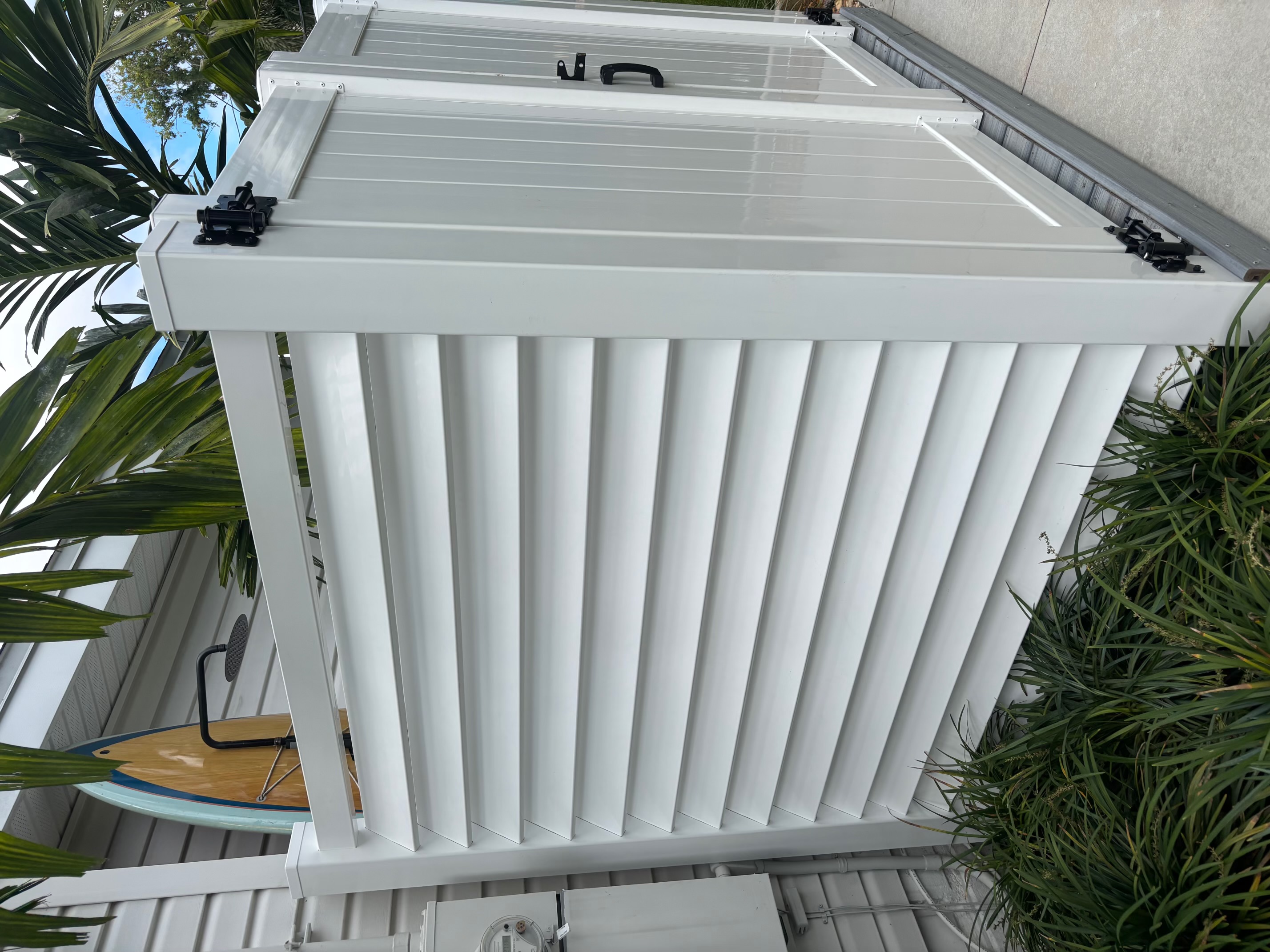 Exterior view of an outdoor white vinyl louvered privacy enclosure with black hardware and surrounding palm foliage