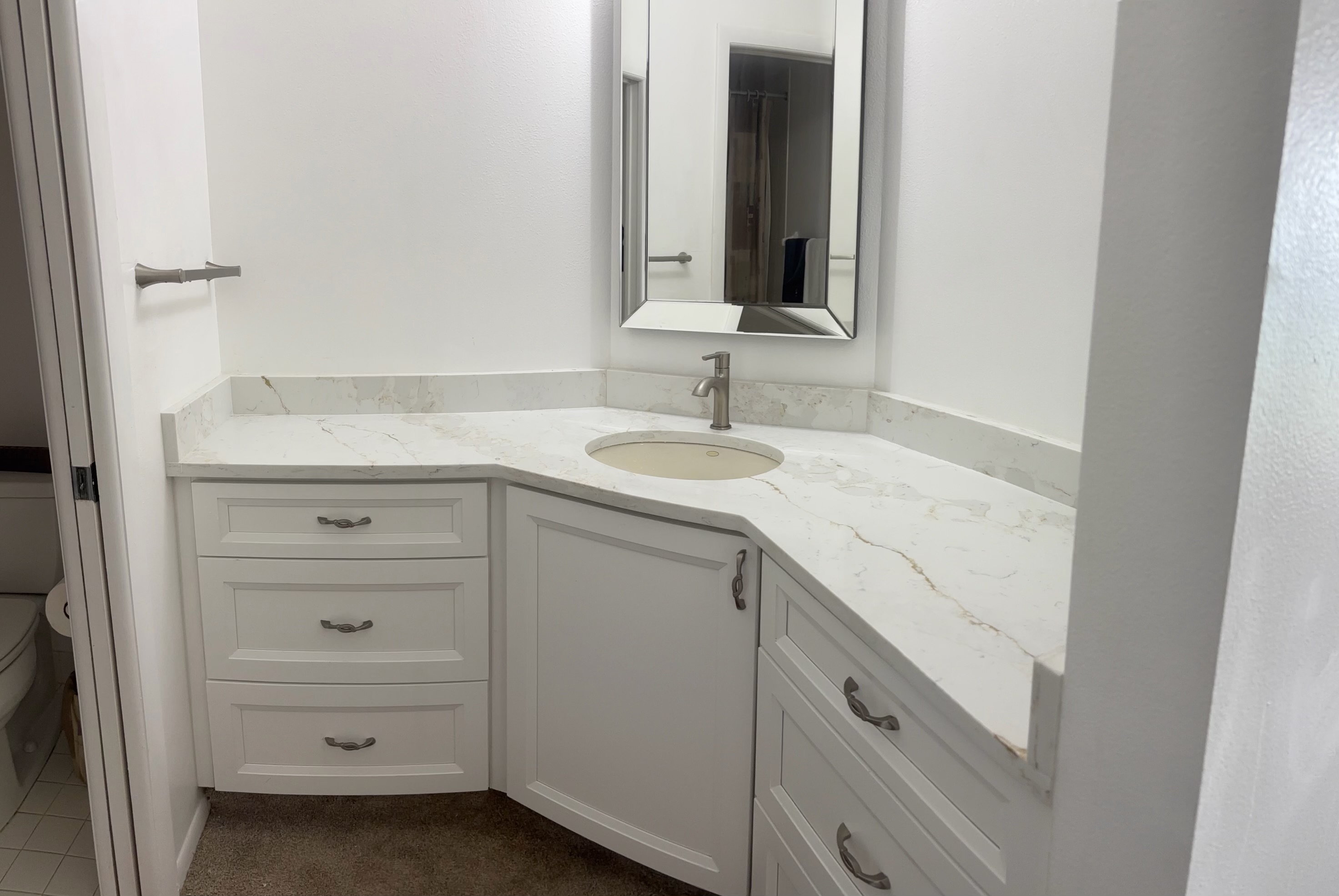 Custom corner bathroom vanity with white shaker drawers and cabinets, marble quartz countertop, and beveled mirror
