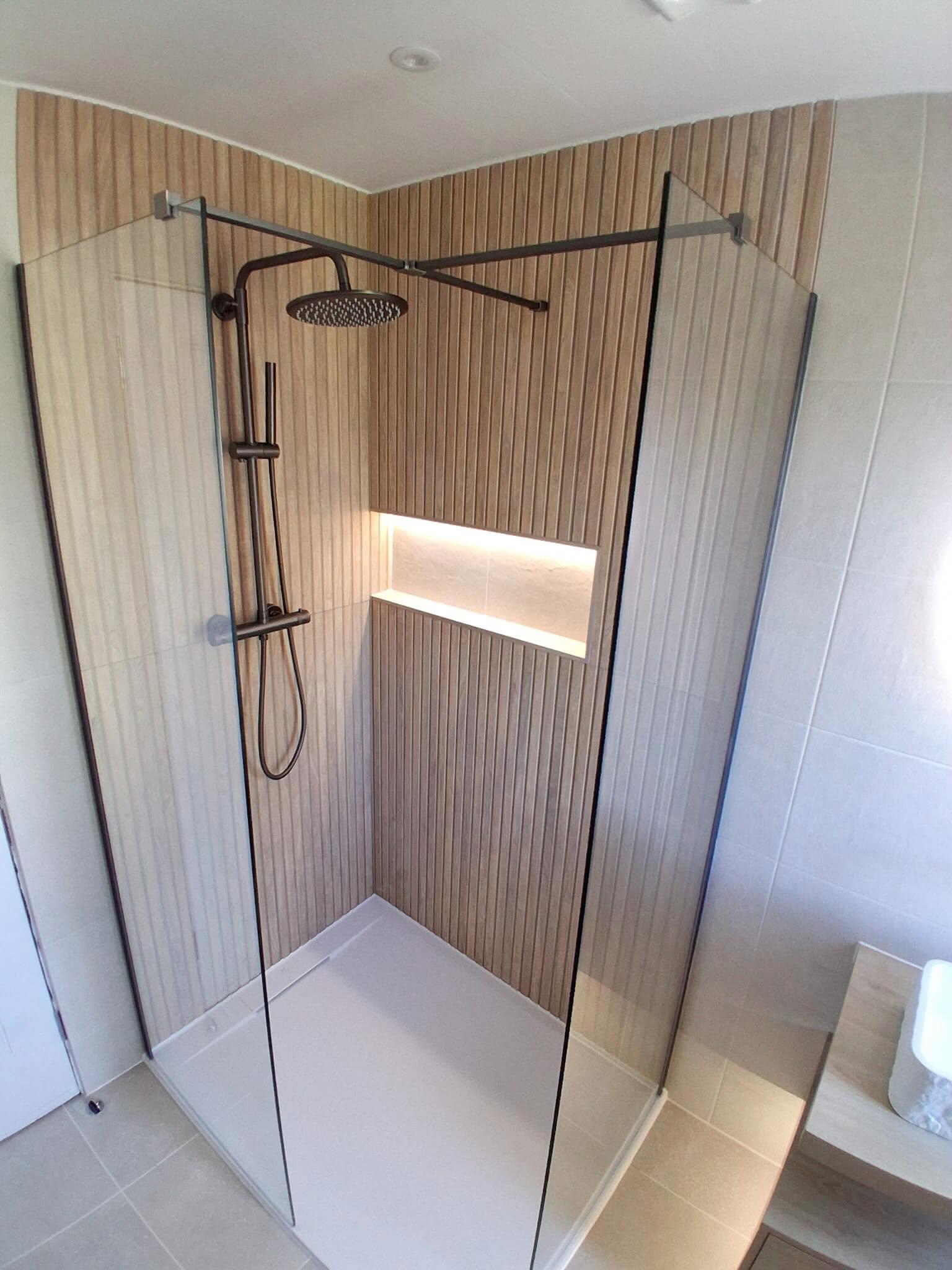 High-angle view of walk-in corner glass shower with vertical oak timber slats, illuminated recessed shelf niche and dark bronze rainfall shower