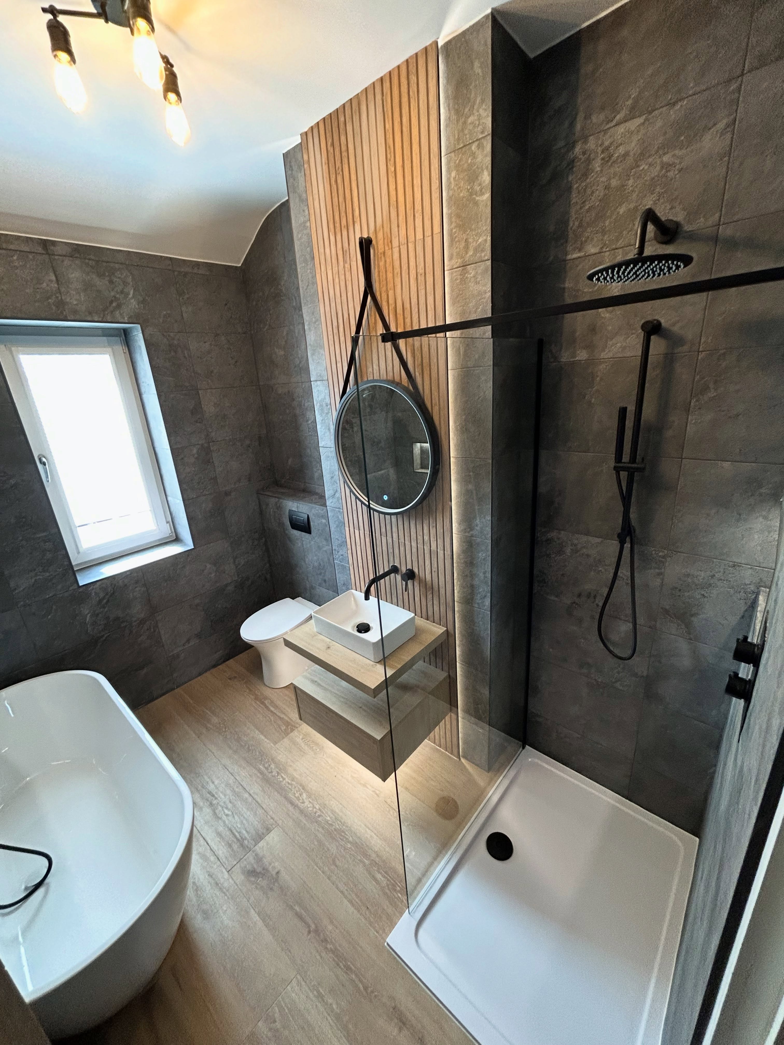 Contemporary master bathroom with charcoal slate wall tiles, vertical wood slat feature column, freestanding bath, walk-in shower and hanging round mirror