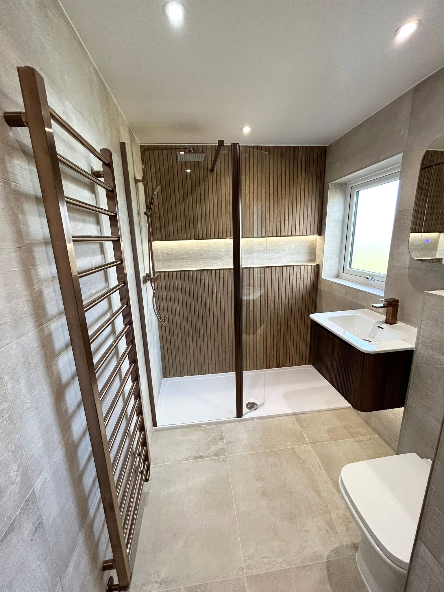 Spacious wet room with full-width wood slat shower wall, horizontal warm LED recessed niche, oversized bronze heated towel rail and fluted vanity