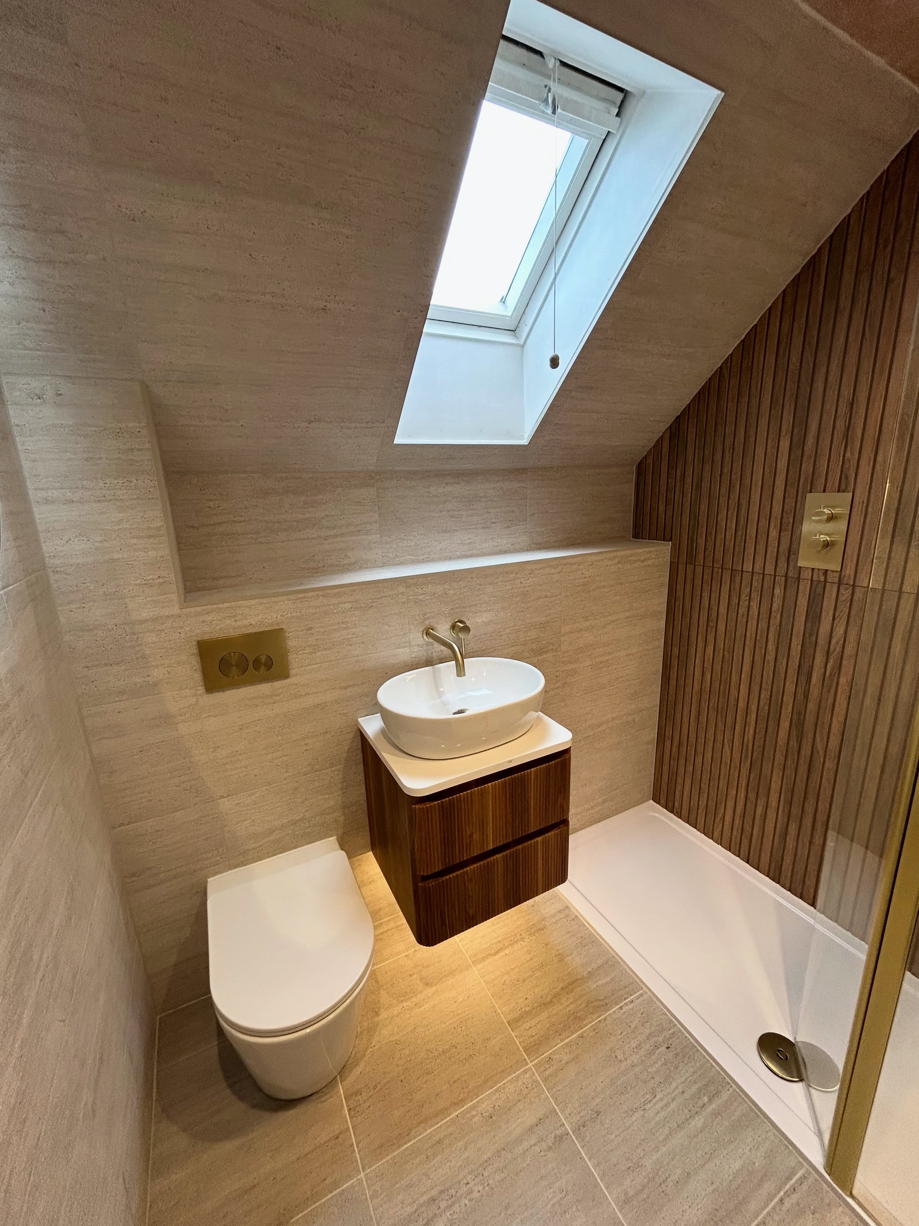 Vaulted ceiling loft bathroom with roof window, travertine tiling, vertical wood slatted shower wall, floating oak vanity and brushed brass fixtures
