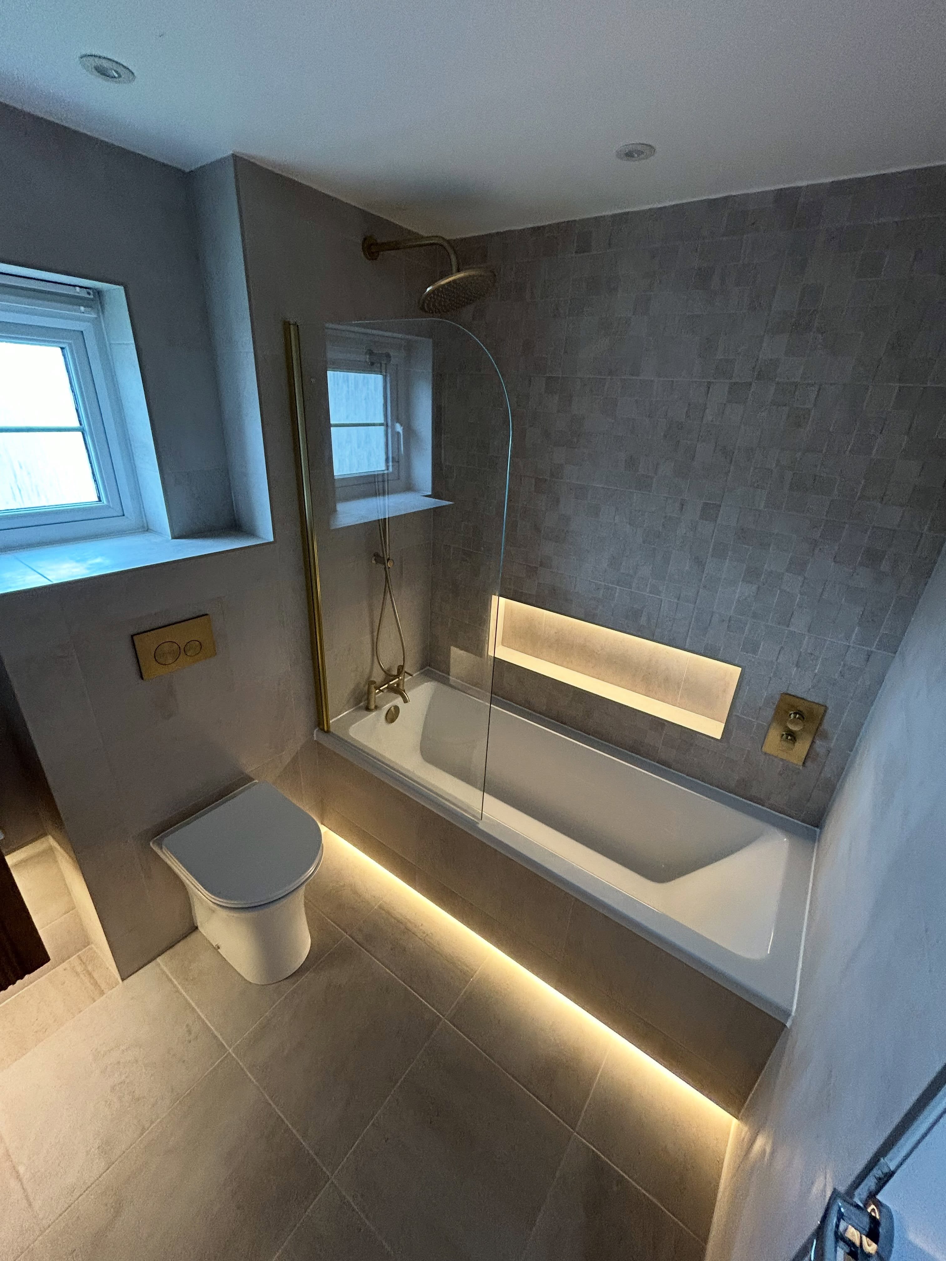 Modern fitted bath with curved glass screen, warm under-bath LED plinth lighting, illuminated recessed shower niche and brushed brass tapware