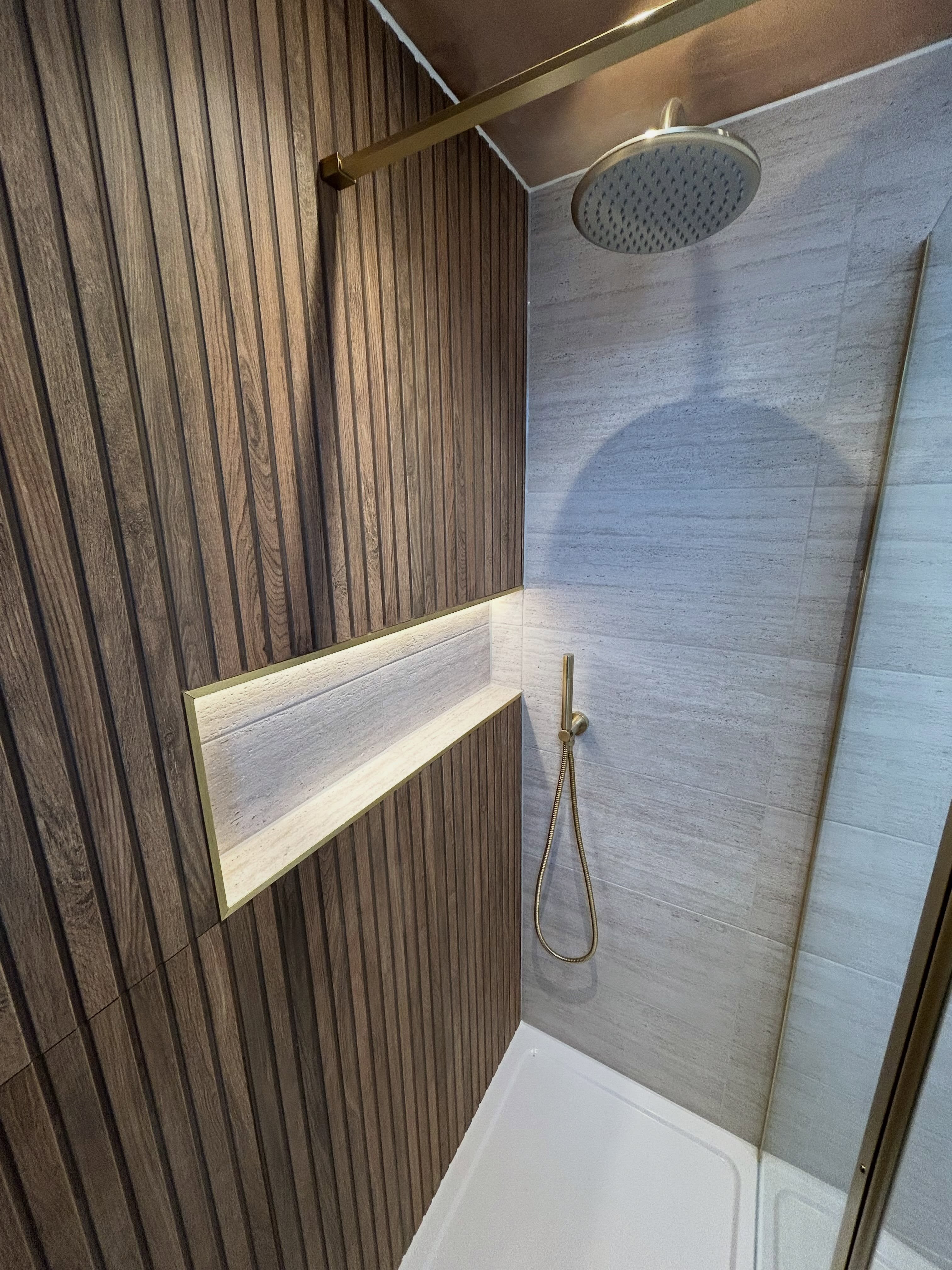 Close-up of walk-in shower with fluted wood panelling, brushed gold shower arm and head, and warm LED illuminated tile niche