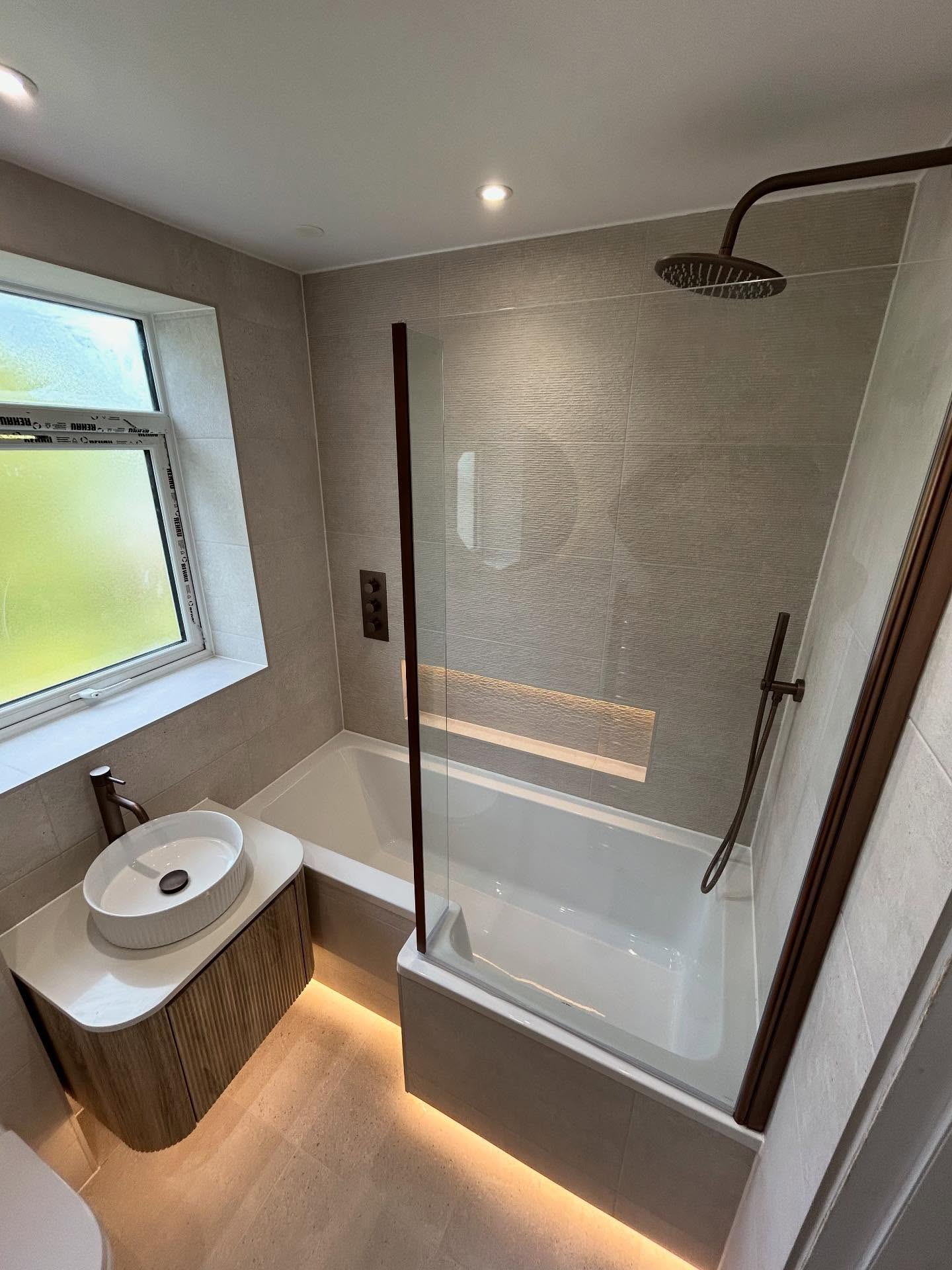 Modern fitted shower bath combination with textured tile feature wall, illuminated recessed niche, concealed bath plinth underglow, and fluted vanity with round basin