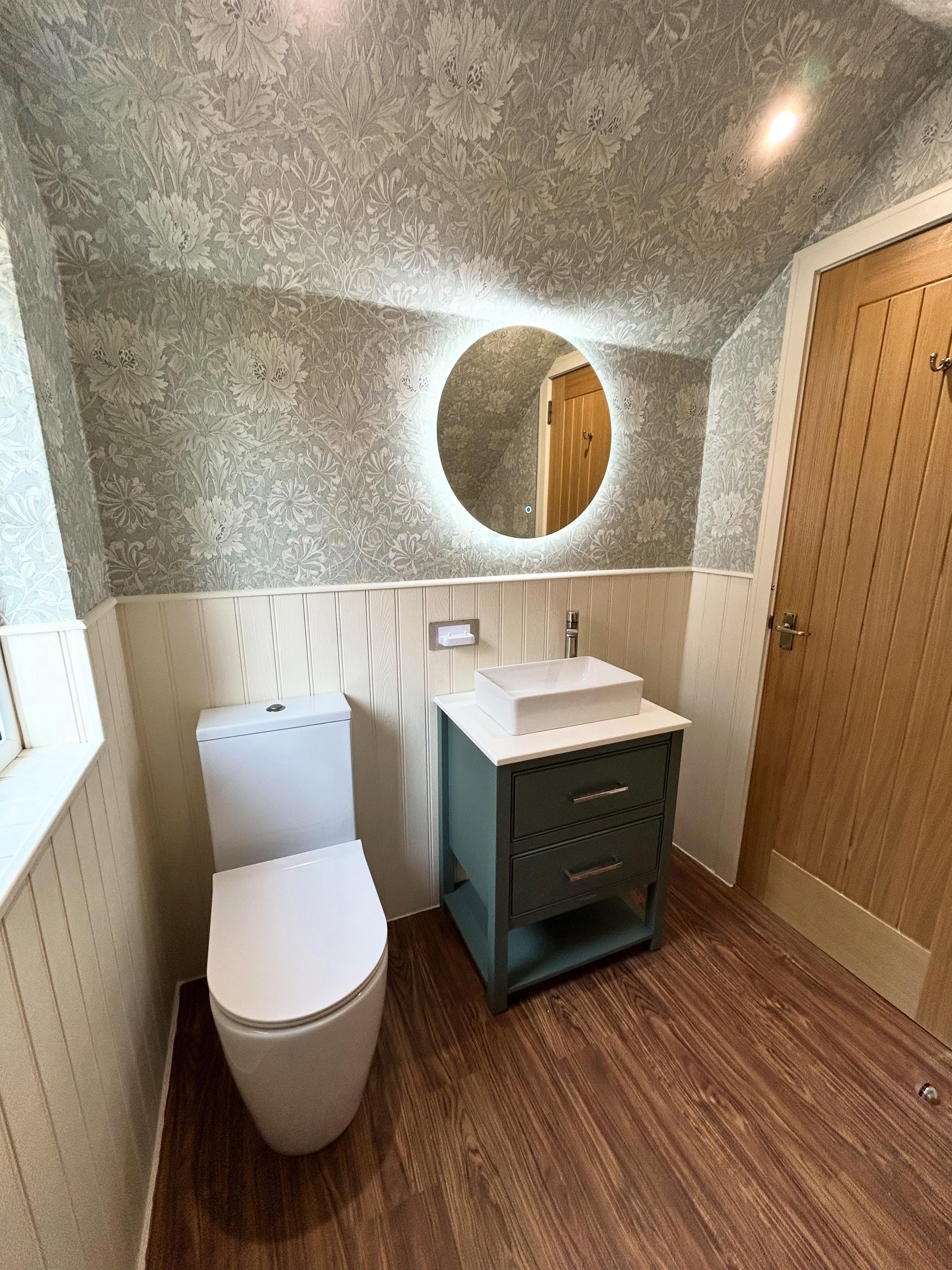 Attic cloakroom with botanical floral wallpaper, vertical tongue-and-groove wainscot panelling, sage green shaker vanity and round halo mirror