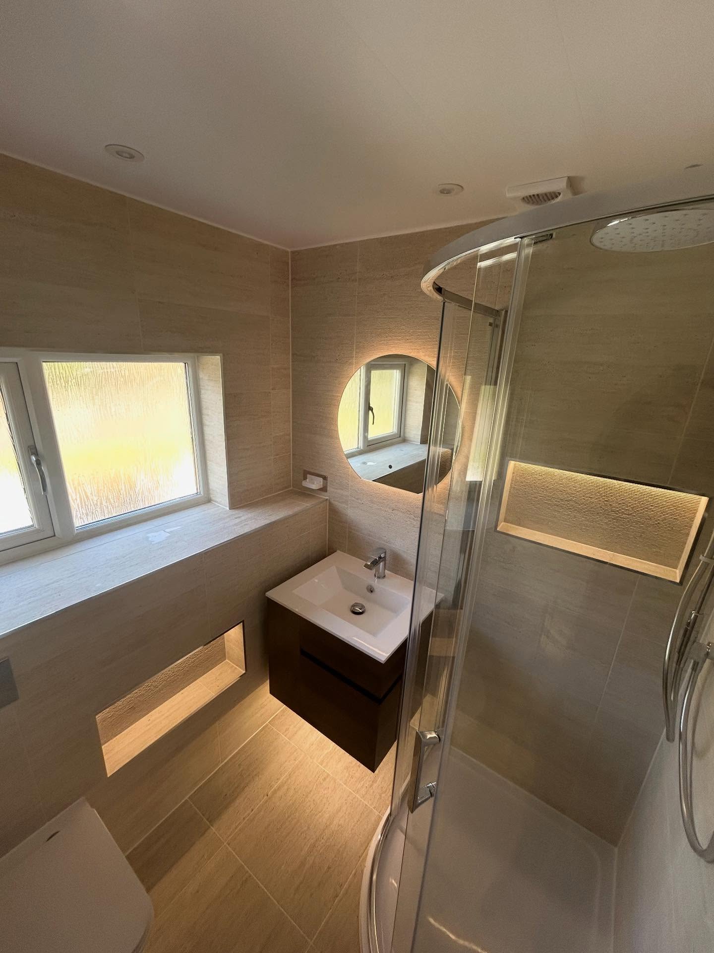 Compact luxury travertine ensuite with curved quadrant glass shower, textured illuminated shower niche, low-level floor niche and halo backlit mirror