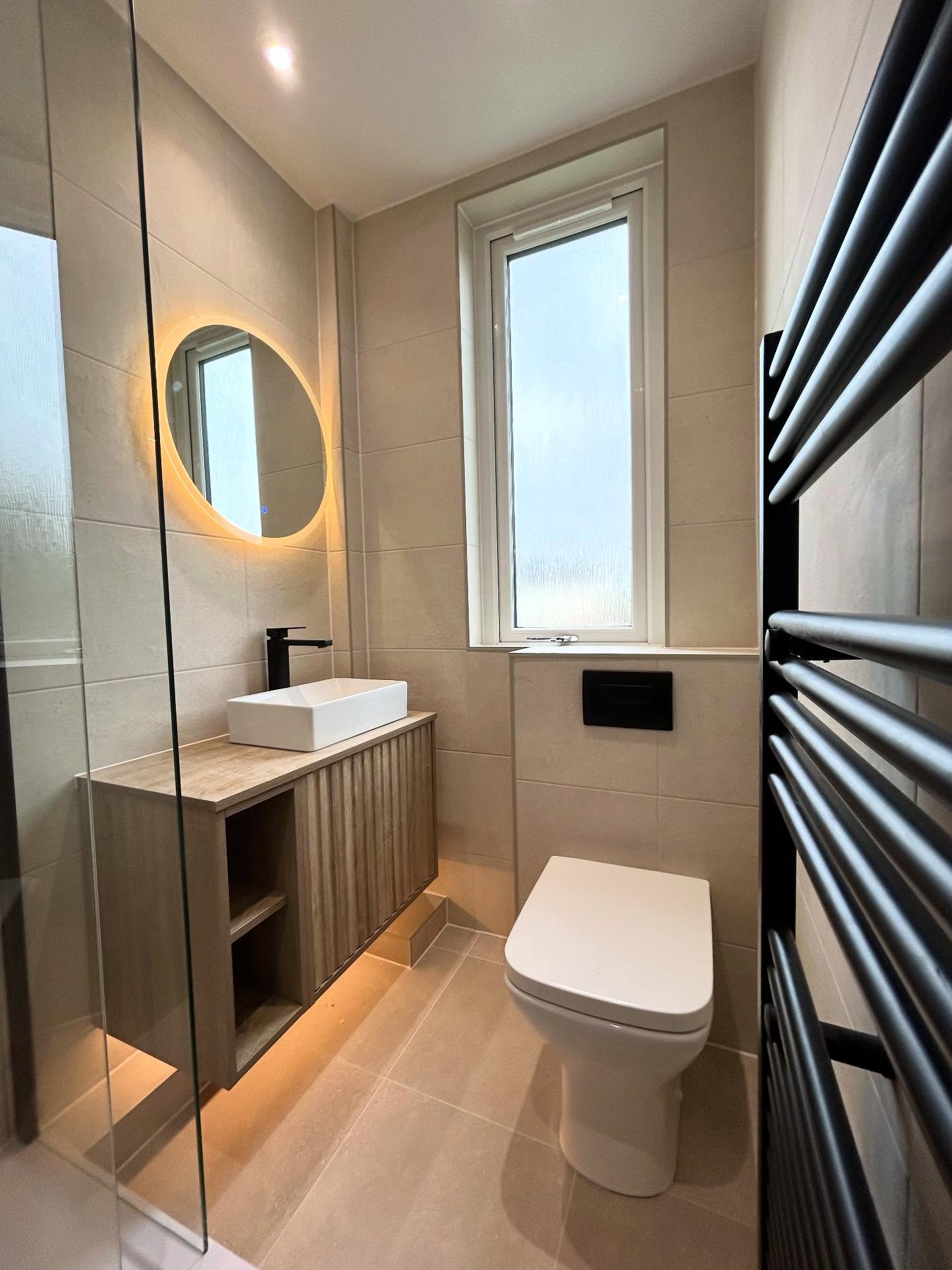 Travertine ensuite bathroom with fluted wood vanity, warm under-cabinet lighting, rectangular countertop basin, backlit round mirror and black towel radiator