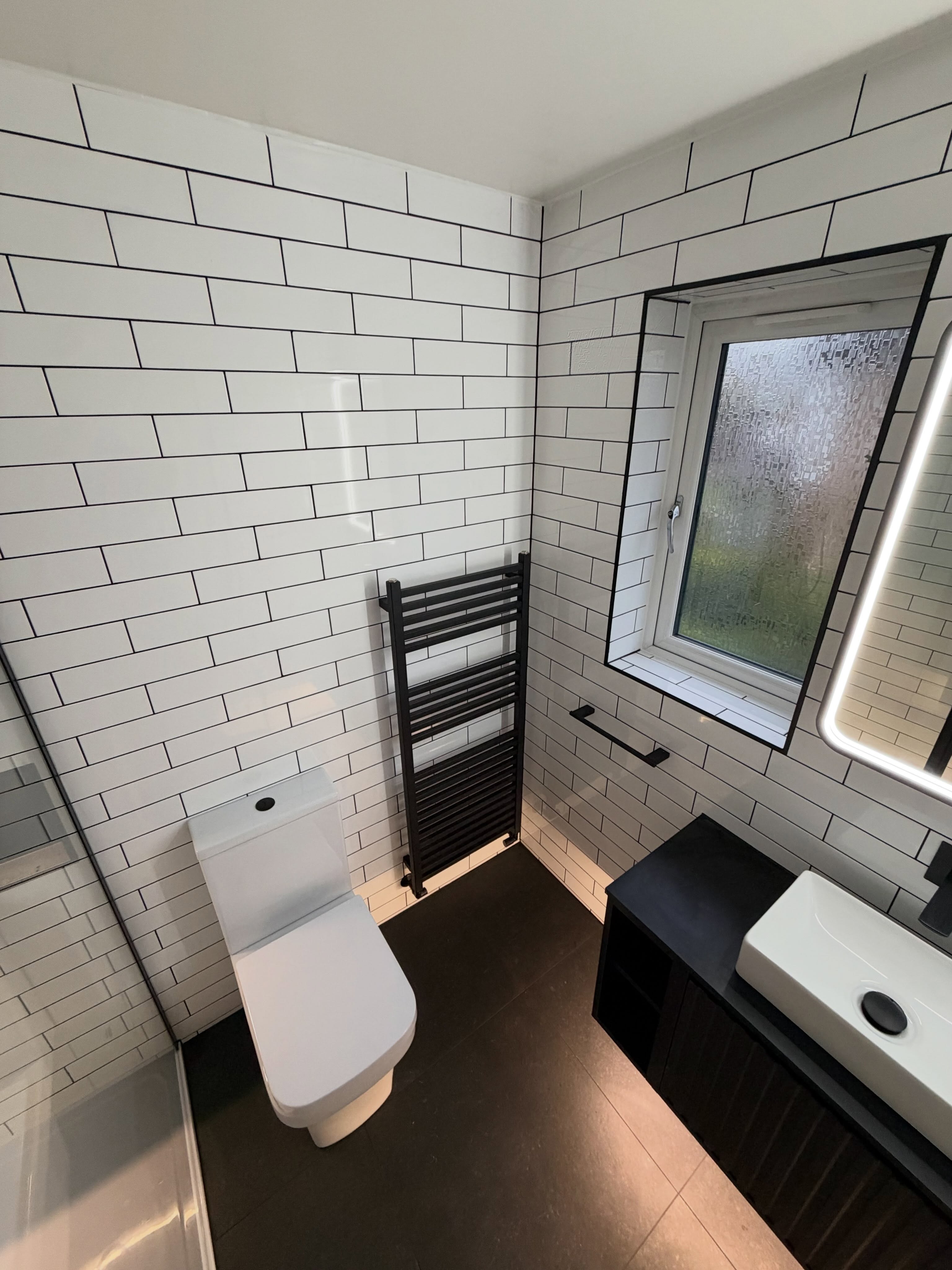 Full bathroom renovation with subway wall tiles, charcoal floor tiling, matte black radiator, illuminated mirror and black vanity unit